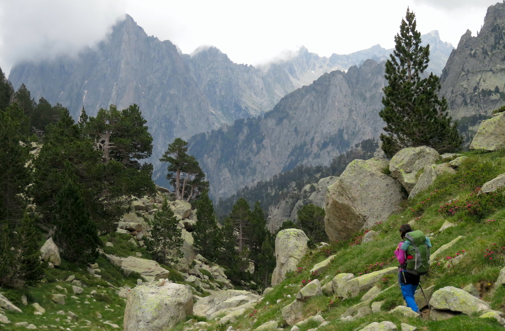 A hiker walking with a trekking backpack along the Carros de Foc trail, with the mountains of the Aigüestortes and Estany de Sant Maurici National Park in the background.